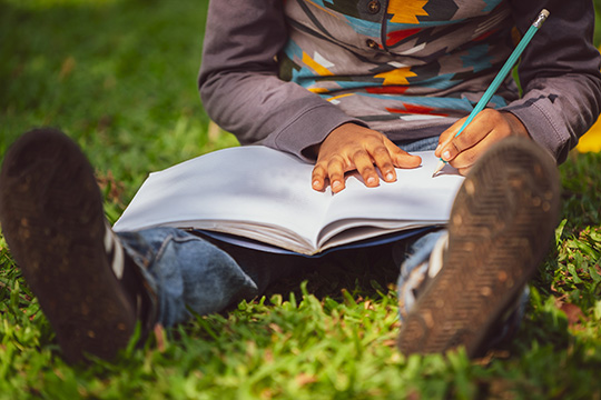 Little child schoolkid writing on a notebook with pencil while sitting on green grass in the park. Kid education concept.