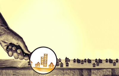 Hand holds the magnifying glass in front of an open newspaper with paper houses. That could mean rent, search, purchase real estate.