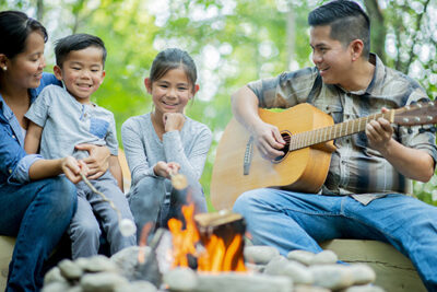 An Asian family is camping outdoors. The father is playing guitar while they sit by the campfire.